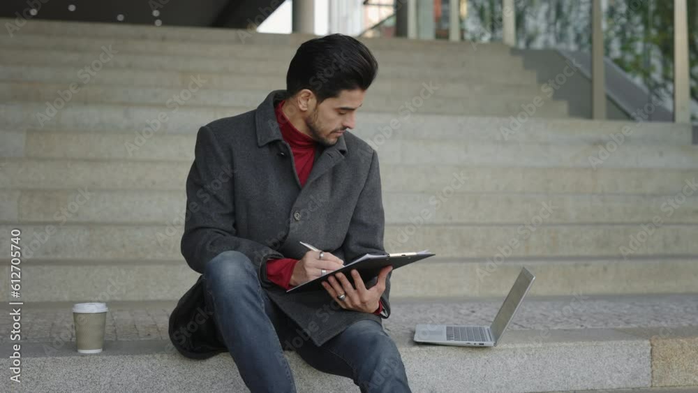 Multi-Ethnic Student Engaged in Laptop Work on University Stairs. A Young Lawyer is Preparing for the Exam. Man looking Through Notes in Notebook. People and Technology Concept Multi-Ethnic Student Engaged in Laptop Work on University Stairs. A Young Lawyer is Preparing for the Exam. Man looking Through Notes in Notebook. People and Technology Concept