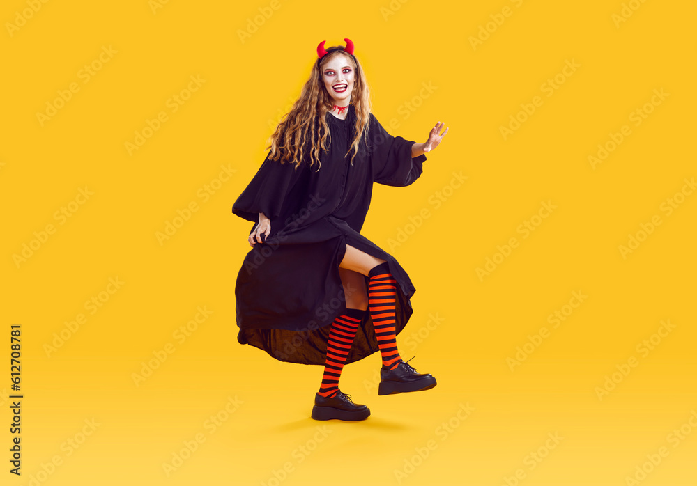 Cheerful young woman in devil costume has fun dancing at Halloween costume party. Woman in long black dress, leggings and red horns is having fun and fooling around on orange background. Full length.