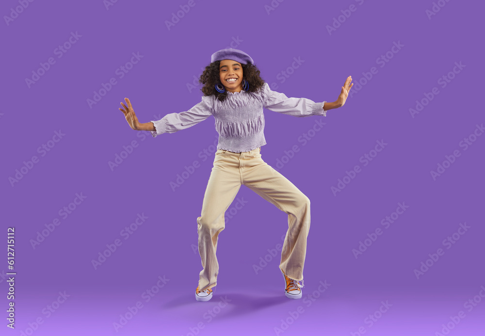 Happy child dressed in cool trendy fit. Full body length of confident girl with curly hair wearing beret hat, blouse and wide leg jeans dancing on purple background. Clothes, kids' fashion concept