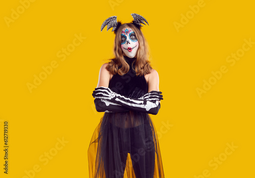 Studio portrait of child in Halloween costume. Teenage girl with sugar skull make up standing with arms folded isolated on yellow background and looking at camera with funny surprised face expression
