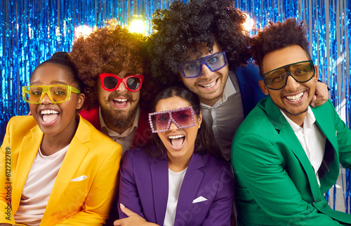 Portrait of a multinational funny group of laughing people dressed in bright costumes, voluminous curly wigs and large glasses on a shiny blue background. Three cute guys and two beautiful girls. photo