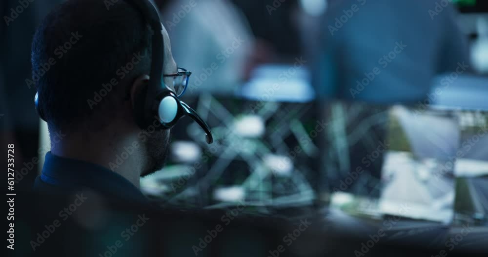 Technical Customer Support Specialist is Talking on a Headset while Working on a Computer in a Call Center Control Room Filled Display Screens and Data Servers. Footage from the Back