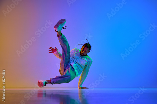 Fototapeta Naklejka Na Ścianę i Meble -  Young guy with dreads, in casual clothes dancing hip-hop, breakdance against gradient multicolored studio background in neon light