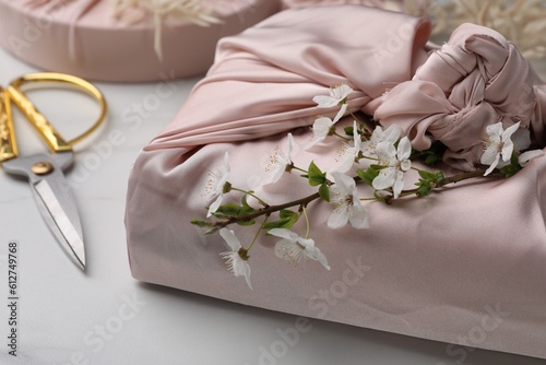 Furoshiki technique. Gift packed in pink fabric, beautiful flowers and scissors on white table, closeup