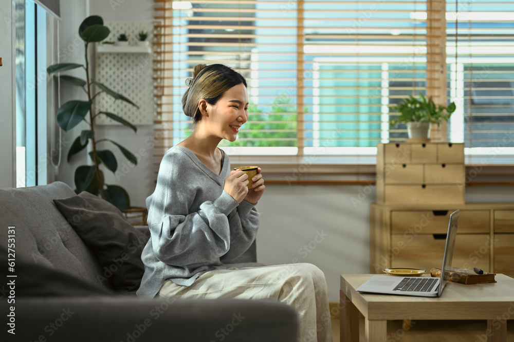Side view of cheerful young woman having video call on laptop and drinking coffee on couch at home Side view of cheerful young woman having video call on laptop and drinking coffee on couch at home