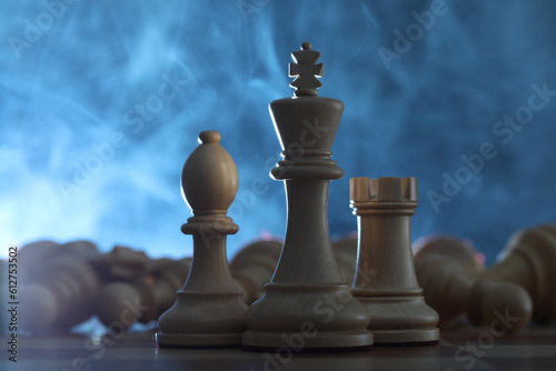 Wooden king  rook and bishop in front of fallen chess pieces on checkerboard against blue background  closeup