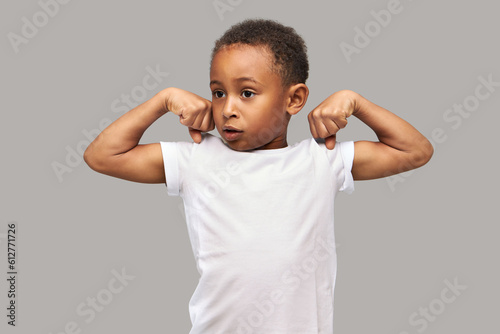 Studio portrait of cute strong muscled little african american kid showing biceps of both arms, looking aside, feeling proud with his strength and endurance, dressed in white mockup t-shirt