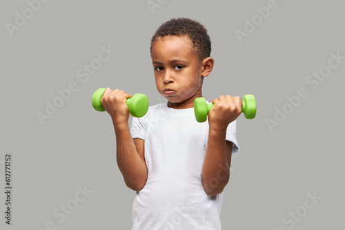 Serious black boy of 8 in white mockup t-shirt training against gray studio background holding green dumbbells, blowing cheeks with exhaustion, keeping fit. Healthy childhood, fitness kids