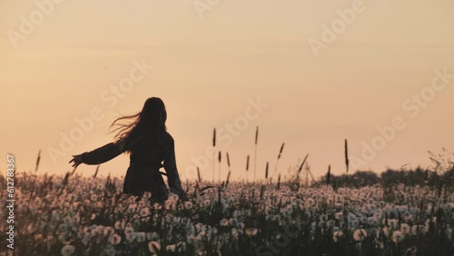 Side view of a happy boho girl running across a field with dry dandelion flowers against the background of a sunset sun. Leisure rest in the evening in the spring. Concept of Peace and Youth