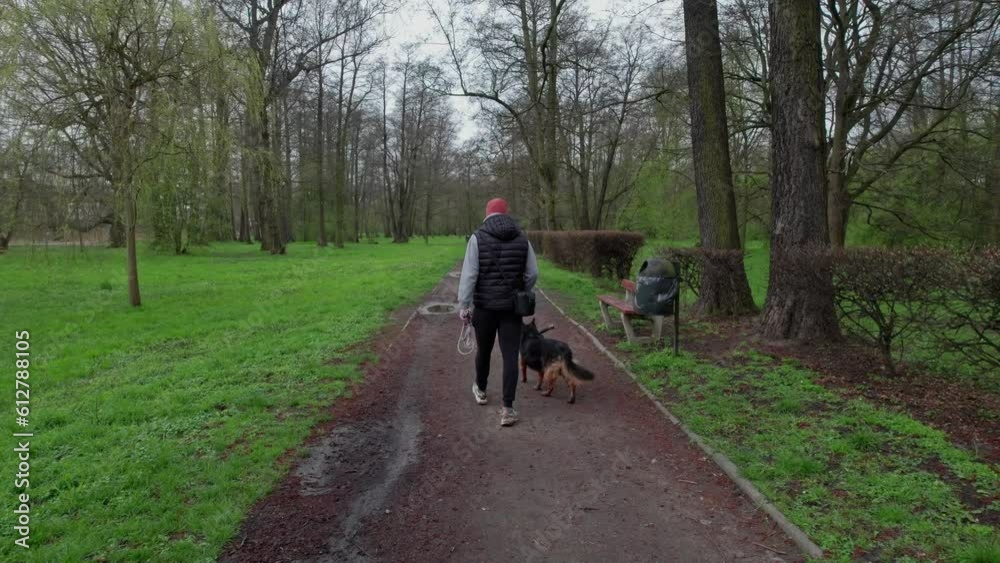In the park on a spring day, a man in a cap and vest strolls with his dog, under a cloudy sky. In the park on a spring day, a man in a cap and vest strolls with his dog, under a cloudy sky.