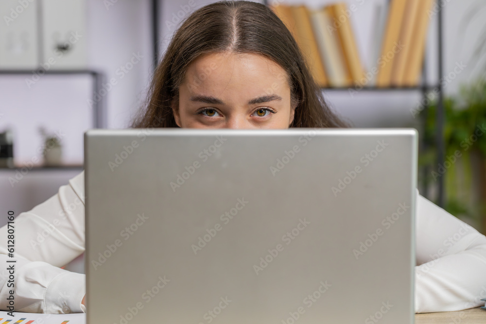 Confident business woman hiding behind laptop computer, looking at camera, spying his colleagues working, peeping. Professional freelancer girl looking from behind computer with cunning eyes glance