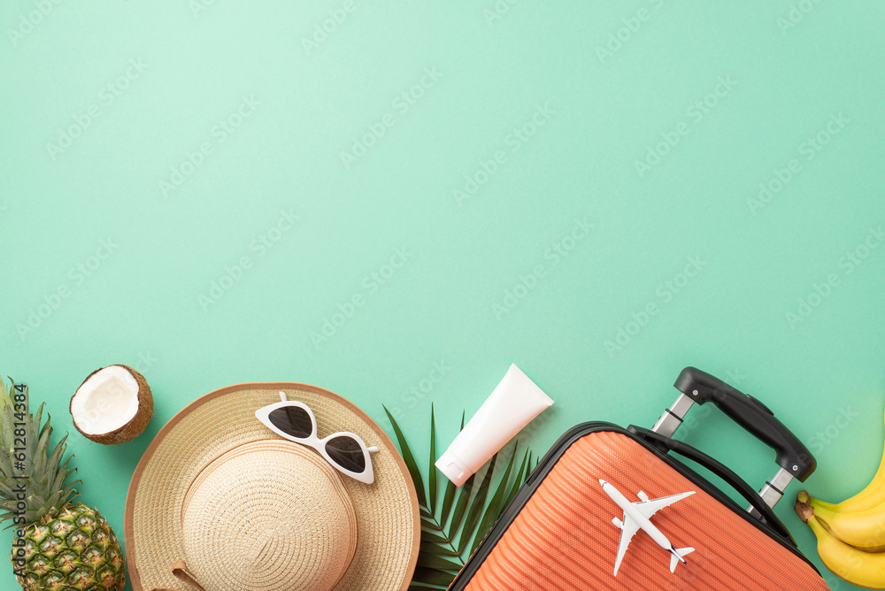 Summer vacation anticipation. Top view of suitcase, small plane replica, beach accessories, glasses, cap, sunscreen tube, palm leaf, exotic fruits on turquoise backdrop with empty area for text or ad Summer vacation anticipation. Top view of suitcase, small plane replica, beach accessories, glasses, cap, sunscreen tube, palm leaf, exotic fruits on turquoise backdrop with empty area for text or ad