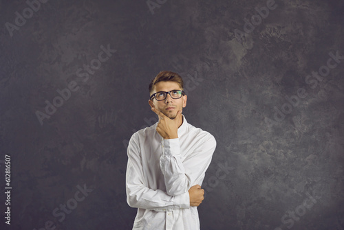 Portrait of serious confused intrigued undecided young man in glasses standing in studio, looking up, holding hand on chin, thinking, doubting, trying to make difficult choice or answer hard question