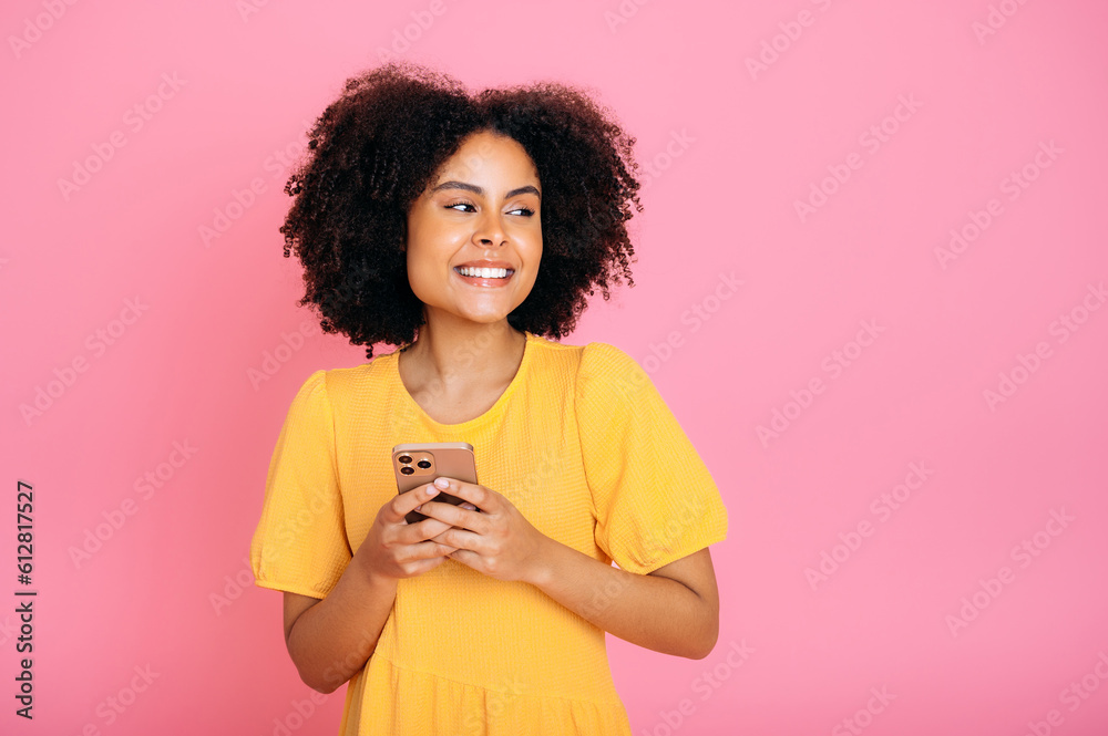 Positive hispanic or brazilian woman, in yellow sundress, using her smartphone, chatting online with friends, browsing internet, texting, looks to the side, smiling, stand on isolated pink background Positive hispanic or brazilian woman, in yellow sundress, using her smartphone, chatting online with friends, browsing internet, texting, looks to the side, smiling, stand on isolated pink background