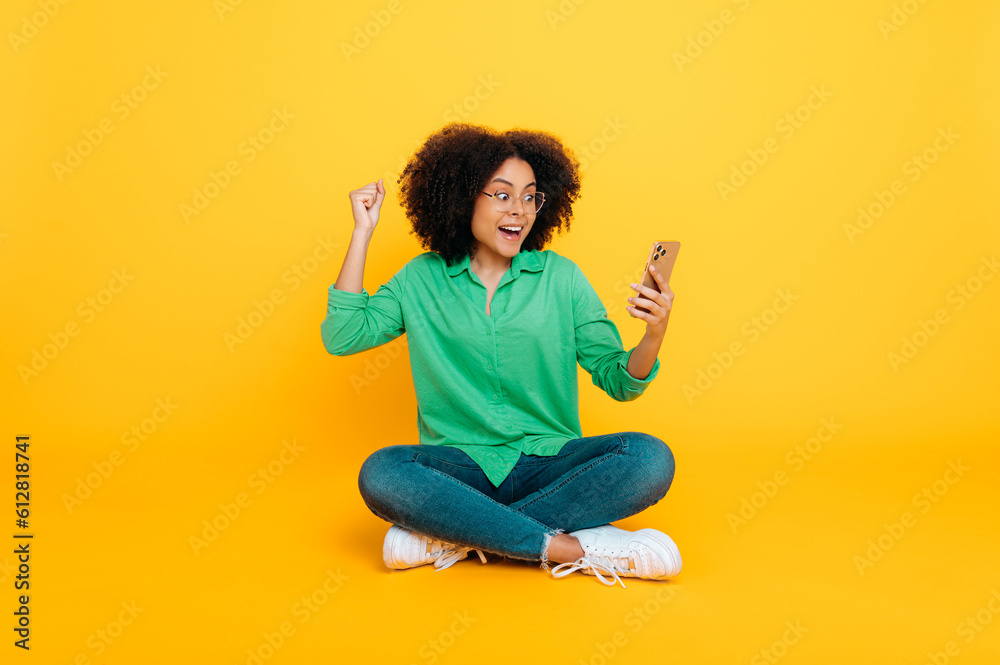 Full length photo of excited happy african american or brazilian curly woman, sits on a yellow background, uses her smartphone, browsing internet, rejoices at the news, gesturing hands, smiles