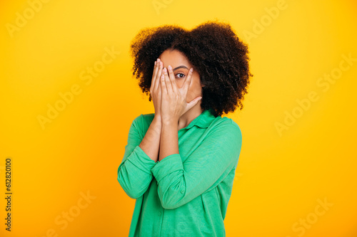 Scary, afraid. Terrified hispanic or brazilian curly haired woman dressed in casual shirt, peeks through fingers, covers face with hands, experiencing scare, shock, stand on isolated yellow background