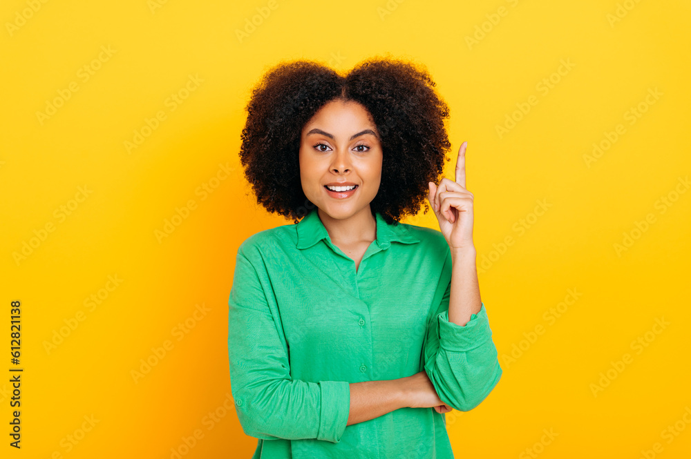 Have idea. Excited curly haired brazilian or hispanic stylish positive woman, in green shirt, looks enthusiastically at the camera, shows IDEA gesture with finger, stands on isolated yellow background Have idea. Excited curly haired brazilian or hispanic stylish positive woman, in green shirt, looks enthusiastically at the camera, shows IDEA gesture with finger, stands on isolated yellow background