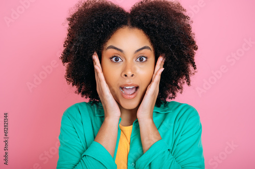 Amazed emotional african american or brazilian curly haired beautiful woman in green shirt, looking at camera in surprise, holding palms near cheeks and opening her mouth, isolated pink background © Kateryna