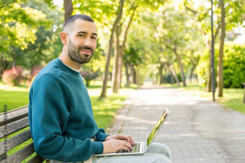 Young bearded man smiling, sitting on a park bench with a laptop