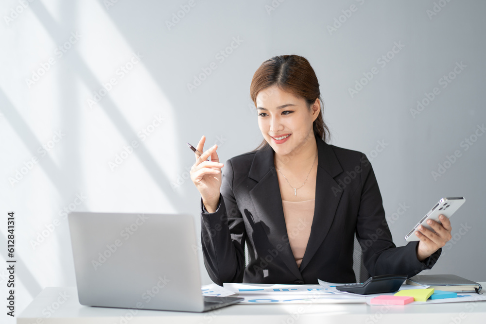 Professional young Asian office worker working on her project with laptop and using her phone at the same time. Professional young Asian office worker working on her project with laptop and using her phone at the same time.