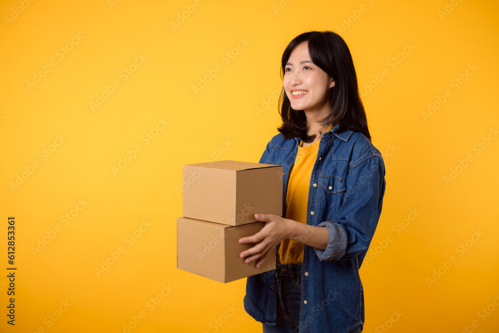 Portrait asian young woman wearing yellow t-shirt and denim shirt holding parcel box isolated on yellow studio background, Delivery courier and shipping service concept. Portrait asian young woman wearing yellow t-shirt and denim shirt holding parcel box isolated on yellow studio background, Delivery courier and shipping service concept.