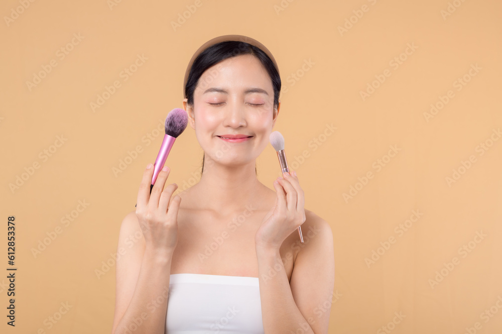 Naklejka premium Portrait happy young asian woman with natural make up face holding cosmetic skin powder blusher isolated on beige background. Female apply skincare brush treatment. beauty product cosmetology concept.