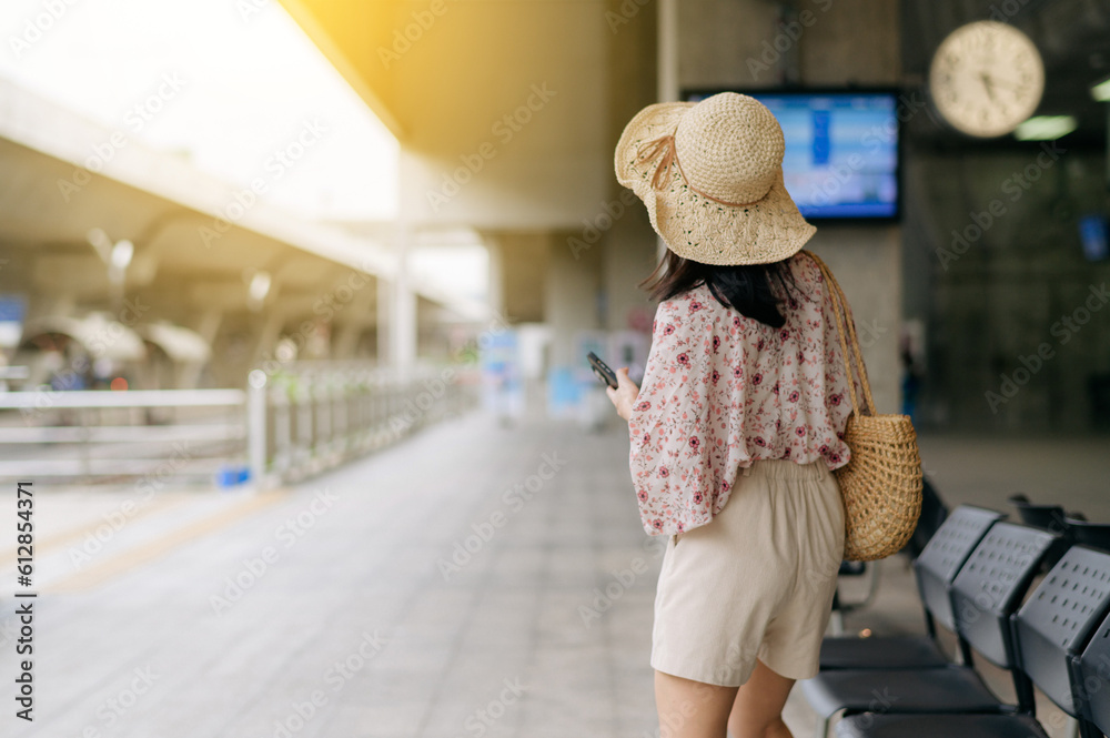 young asian woman traveler with weaving basket using a mobile phone and waiting for train in train station. Journey trip lifestyle, world travel explorer or Asia summer tourism concept.