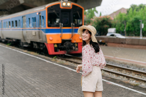 young asian woman traveler with weaving basket looking back happy smiling to a camera beside train coming background. Journey trip lifestyle, world travel explorer or Asia summer tourism concept.