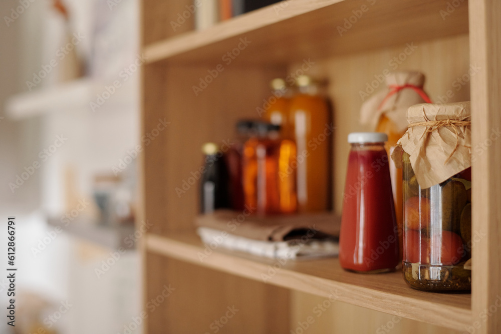 Focus on group of jars with homemade ketchup, pickled tomatoes and other food products standing on wooden shelf in the kitchen