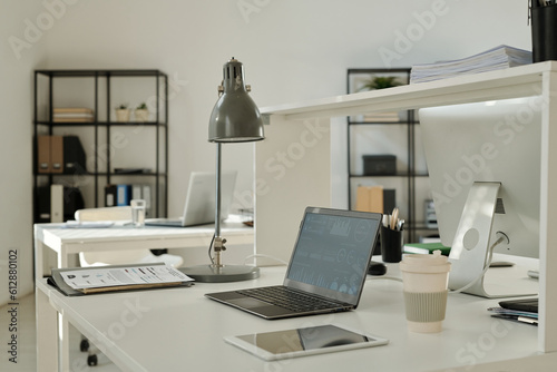 Financial graphic data on screen of laptop standing on desk among paper documents, lamp, tablet and cup of coffee in spacious office