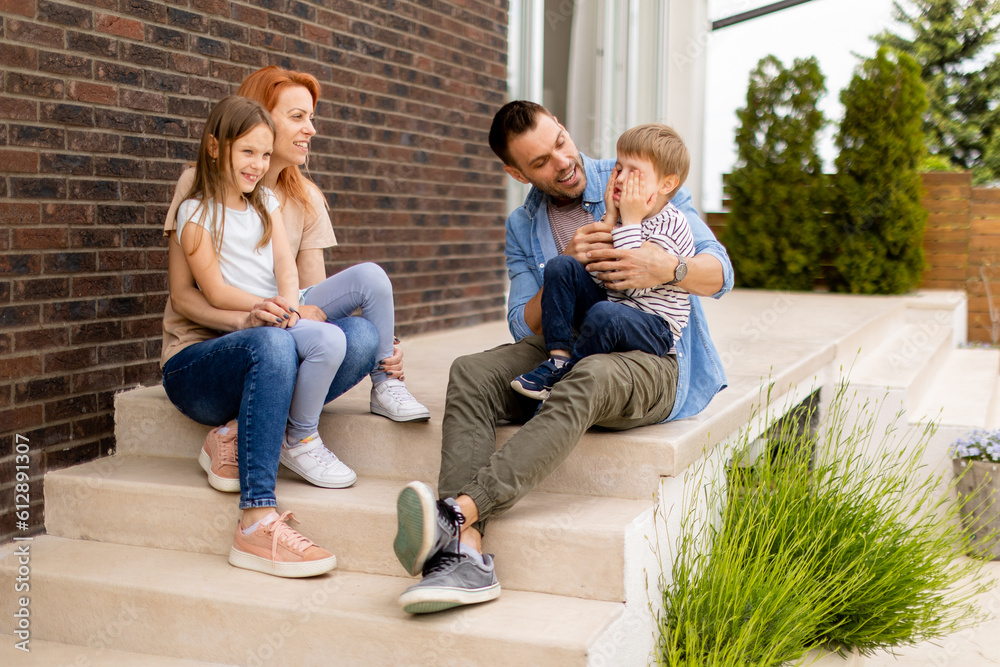 Family with a mother, father, son and daughter sitting outside on the steps of a front porch of a brick house Family with a mother, father, son and daughter sitting outside on the steps of a front porch of a brick house