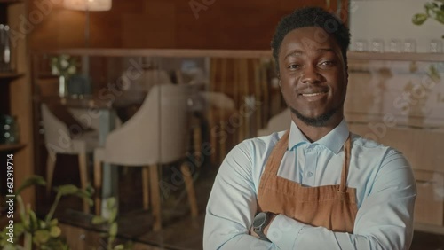 Tilt up portrait of young confident African American waiter posing for camera with hands folded working in posh expensive restaurant