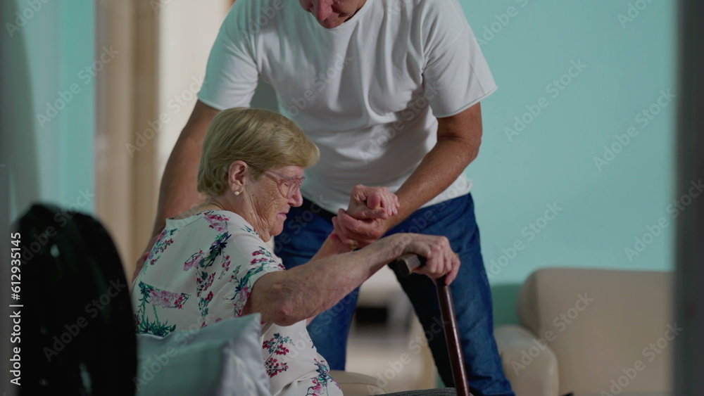Obraz premium Assisting Elderly woman at home, candid authentic scene of adult son helping senior mother tosit on sofa, older woman holding on into stick grip