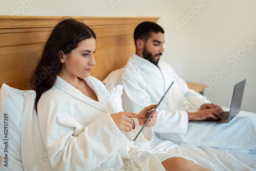 Young Spouses Websurfing With Laptop And Tablet Computers In Bedroom