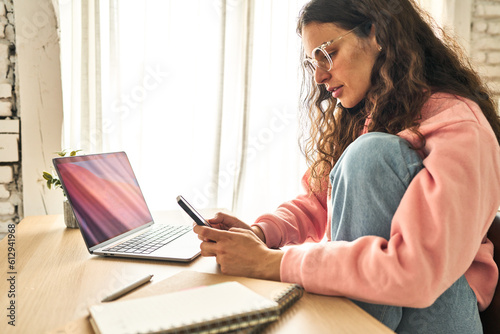 A young woman, focused and productive, working at her home desk with a laptop, researching on her mobile phone.