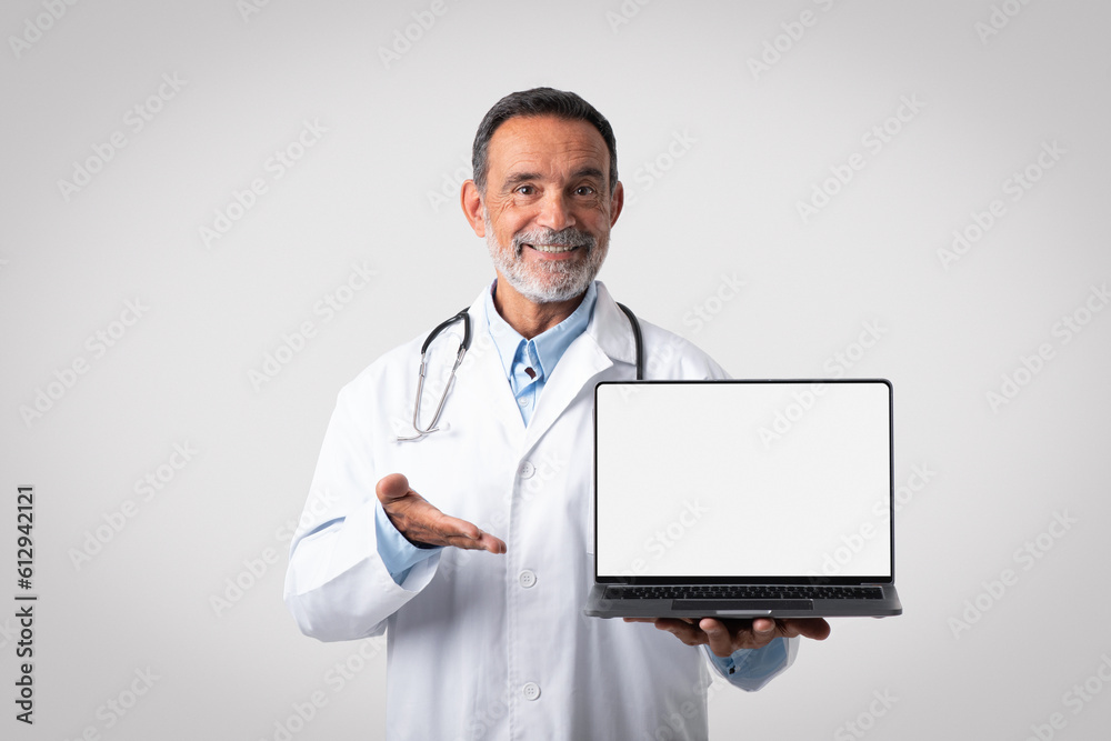 Happy caucasian mature man doctor in white coat point hand on laptop with blank screen