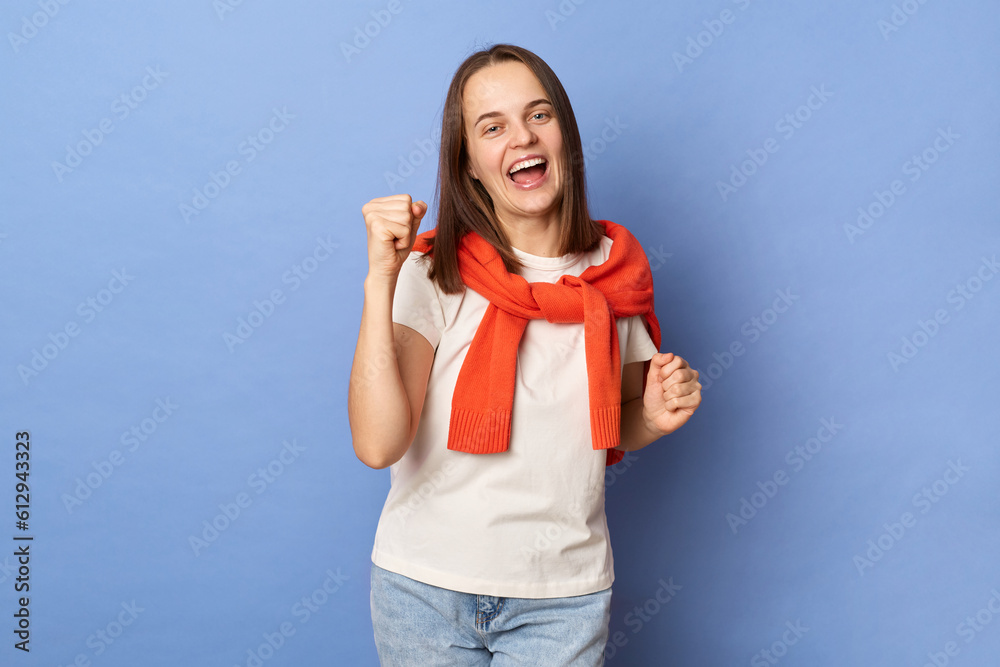 Overjoyed woman wearing white t-shirt and jumper over neck standing isolated over blue background clenched fists celebrating her achievement. Overjoyed woman wearing white t-shirt and jumper over neck standing isolated over blue background clenched fists celebrating her achievement.