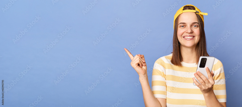 Smiling cheerful teenager girl wearing striped t-shirt and baseball cap standing isolated over blue background holding smart phone pointing at advertisement area.