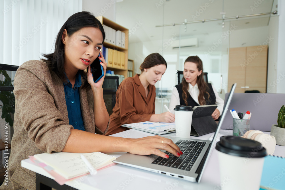 Young businesswoman talking on mobile phone and typing on laptop with her colleagues working in background Young businesswoman talking on mobile phone and typing on laptop with her colleagues working in background