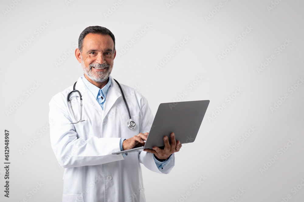 Cheerful caucasian mature man doctor in white coat typing on laptop