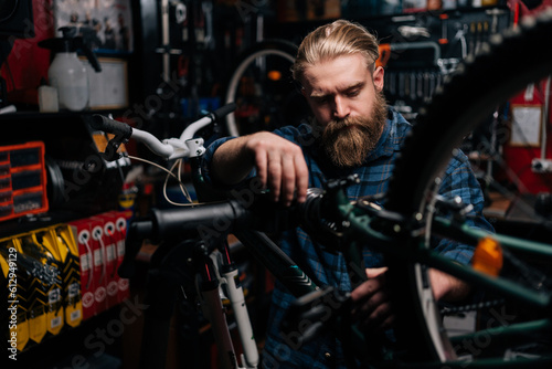 Handsome bearded cycling mechanic male repairing and fixing mountain bicycle standing on bike rack working in bike repair shop with dark interior. Concept of maintenance of bicycle transport.