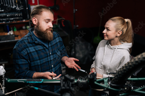 Front view of bearded cycling repairman male having conversation with blonde female client, talking about problem of bicycle detected during diagnostics, in repair shop with dark interior.