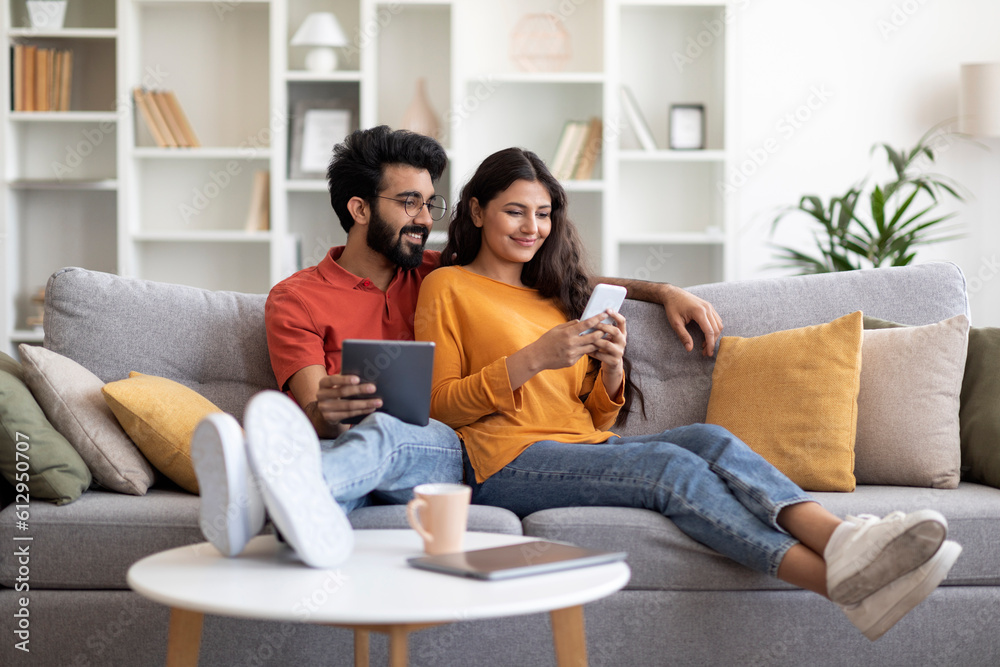 Happy Young Indian Couple Relaxing At Home With Smartphone And Digital Tablet