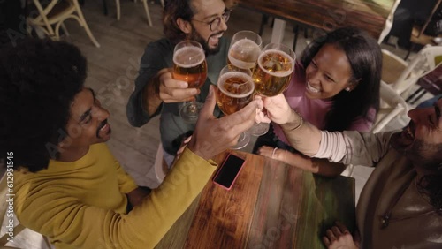 Group of multirracial young people toasting with a pint of beer and celebrating happy hours in a bar indoors. Friends drink alcohol together having fun and smiling. Cheers of gathering latin students