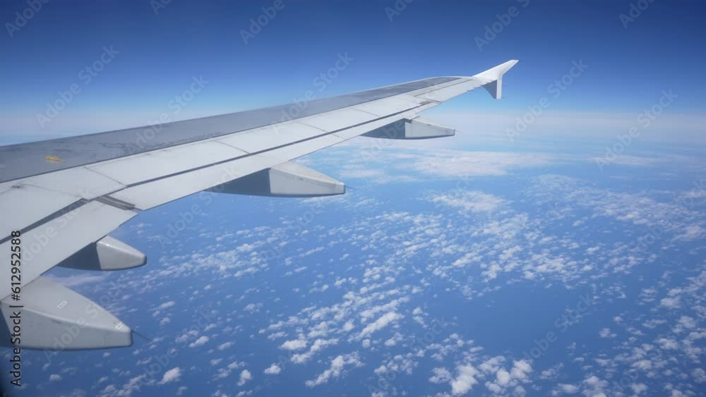 Closeup view through window on wing of airplane flying above white clouds on background of blue sky on sunny day, slow motion. Slowly moving fluffy clouds from airplane window during flight. Closeup view through window on wing of airplane flying above white clouds on background of blue sky on sunny day, slow motion. Slowly moving fluffy clouds from airplane window during flight.