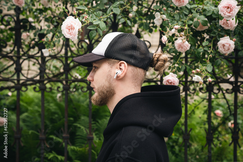 Stylish bearded young man in a cap and headphones against the background of a climbing rose on the fence, the concept of recreation, tourism and the use of gadgets.