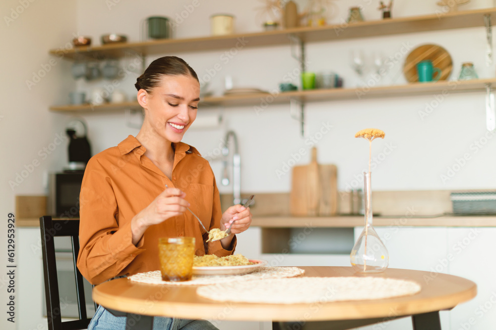 Young caucasian woman eating tasty pasta, having lunch, enjoying delicious homemade spaghetti, sitting in cozy kitchen