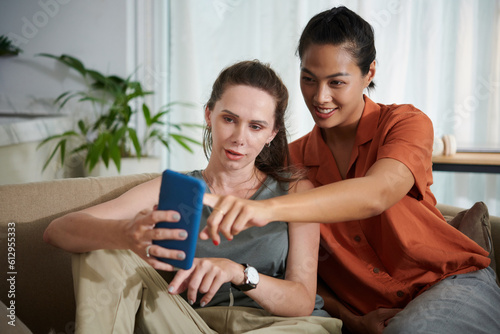 Two friends using smartphone together while sitting on sofa at home