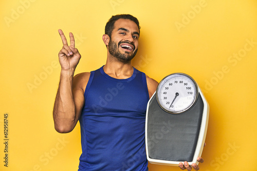 Fit young Latino man with scale on yellow background, fitness concept, joyful and carefree showing a peace symbol with fingers.