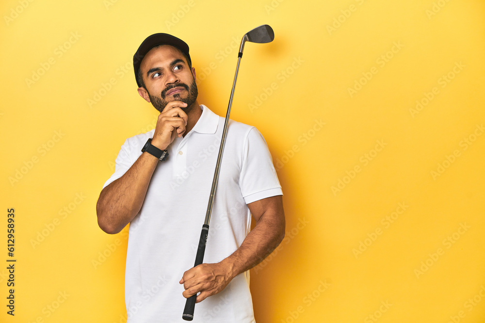 Young Latino golfer with club and cap on a yellow studio background, looking sideways with doubtful and skeptical expression. Young Latino golfer with club and cap on a yellow studio background, looking sideways with doubtful and skeptical expression.